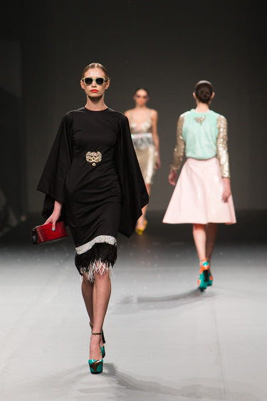 Fashion models walking on a runway during a designer show, featuring elegant and stylish outfits, including a black dress with embellishments and colorful high heels.